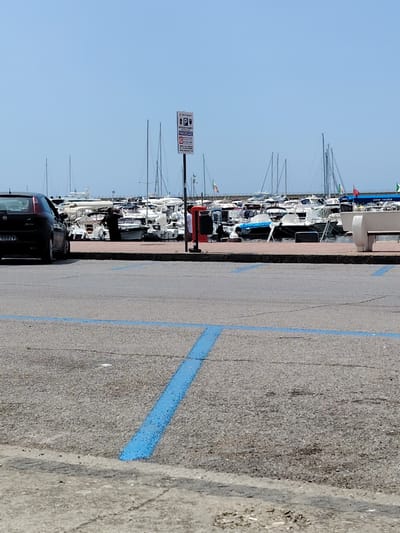 This media file depicts a daytime scene in a marina, likely in Agropoli, Italy, under a clear blue sky. The foreground features a paved parking lot with blue painted lines indicating parking spaces. In the mid-ground, a black car is parked on the left. Beyond the parking lot, a waterfront promenade is visible, lined with numerous docked sailboats and motorboats. Several people are present near the boats, with one individual in dark clothing standing and appearing to look out towards the water or the boats.

A prominent sign with "P ARCHEGGIO" is visible, indicating a parking area, and displays symbols for parking and potentially disabled access. The boats vary in size and type, suggesting a busy marina. The weather appears to be fair and sunny, typical of a pleasant day. The overall atmosphere is calm and suggestive of a recreational area or a place of commerce related to boating. No specific emotional tone is overtly conveyed, but the scene suggests a functional and possibly leisurely setting.