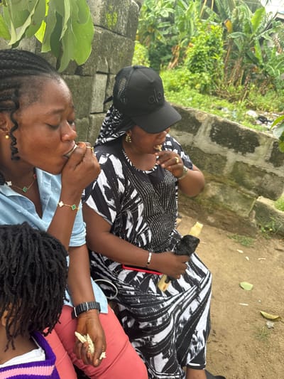 This image captures a candid moment of three individuals, likely women and a child, seated outdoors in Ganyingbo, Nigeria. The background suggests a somewhat rustic environment with a stone wall and lush green vegetation, possibly indicating a residential or communal area within the city.
In the foreground, a woman with braided hair, wearing a light blue shirt and adorned with gold earrings and a clover-themed necklace and bracelet, is seen bringing a small, white object to her mouth. Her eyes are closed, suggesting she is savoring the item. Next to her, another woman is wearing a black and white patterned dress and a black baseball cap. She is actively eating, holding a piece of food in her right hand and looking down at it. Her left hand is also engaged with food. A third person, likely a child, is partially visible on the left, wearing a purple and white striped garment. Their back is to the camera, and their hair is styled in dreadlocks.
The individuals appear to be engaged in eating or drinking, creating a relaxed and informal scene. The overall atmosphere is one of everyday life in a Nigerian setting.