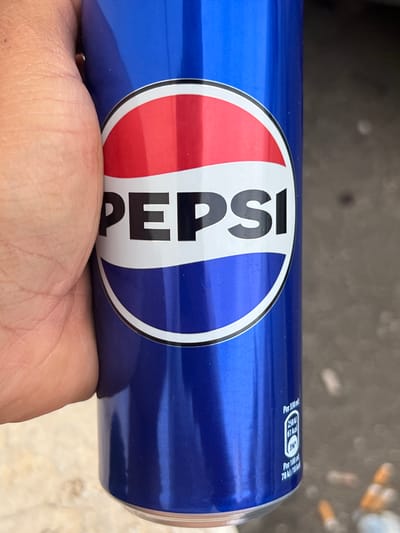 A human hand, displaying visible skin creases and a light skin tone, firmly grips a glossy blue, cylindrical Pepsi can from the left side. The can prominently features the classic Pepsi logo: a white circle outlined in black, horizontally bisected by a white wavy band. The upper segment of the circle is red, and the lower segment is dark blue. The word "PEPSI" is printed in black capital letters across the central white band. On the lower right side of the can, vertically oriented white text against the blue background indicates nutritional information: "Per 330 ml: 258 kJ / 61 kcal" and "Per 100 ml: 78 kJ / 18 kcal." The background is out of focus, depicting an outdoor ground surface. To the right of the can, the ground appears dark grey or brown, with several blurred, small, cylindrical objects, resembling cigarette butts, scattered across it. Directly beneath the can, the ground transitions to a lighter, possibly beige or tan, paved surface. The image contains no discernible features or metadata to confirm a specific geographical location, such as Ontinyent, Spain.
