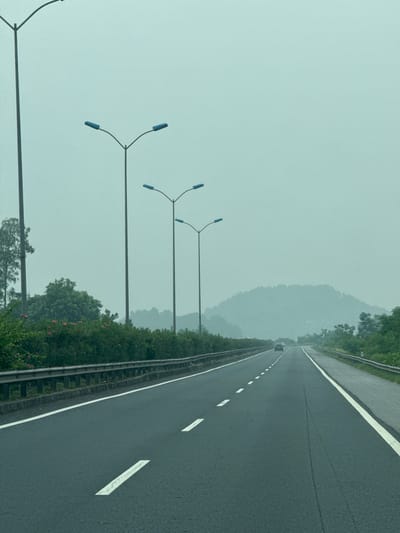The image shows a highway in the city of Tân Thanh, Vietnam, on a foggy day. The road is paved and has white lane markings. On the left side of the road, there is a row of trees and bushes, as well as several tall lampposts with blue lights. On the right side of the road, there is a smaller lane and more greenery. In the distance, there are mountains covered in fog. There are a few cars on the road, driving in the same direction as the camera. The overall scene is serene and peaceful, with a sense of quiet isolation.