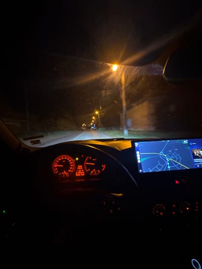 The image shows the interior of a car at night. The dashboard is visible with glowing gauges and a navigation screen displaying a map. Through the windshield, a road stretches ahead, illuminated by streetlights. The location is Mogila, Bulgaria.