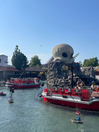 The image depicts an outdoor amusement park scene at LEGØLAND Billund Resort in Billund, Denmark. The central focus is a pirate-themed water ride, with several red and white boats navigating a body of water. In the background, a large skull made of LEGO bricks is prominently featured, perched atop a rocky structure. The sky is clear and blue, indicating a sunny day. People are visible in the boats, enjoying the ride. The environment includes trees, buildings, and a roller coaster track. The overall atmosphere suggests a recreational setting.