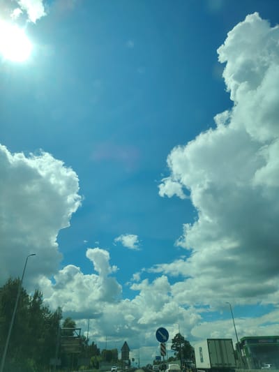 The image depicts a bright, sunny day with a vivid blue sky filled with fluffy white clouds. The sun is visible in the upper left corner, casting a bright light. The perspective suggests a view from a vehicle, likely on a road, as indicated by the presence of traffic signs, streetlights, and other vehicles in the lower portion of the image. Specifically, a white truck with a blue logo is visible, as is a green structure with the word "АПТЕКА" (Apteka, meaning Pharmacy) on it. Trees and foliage line the side of the road. The presence of traffic and road infrastructure strongly suggests an urban or suburban environment. Based on the provided context, this scene is located on Peremohy Street in Ukraine.