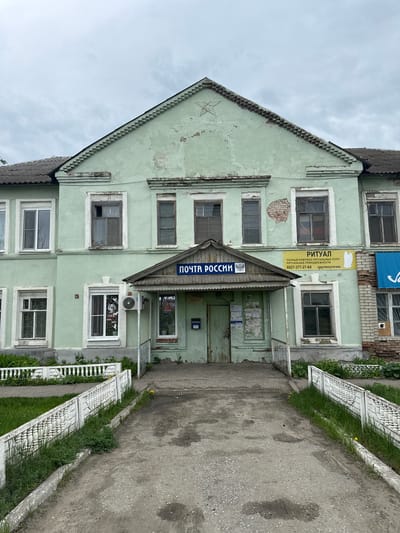 The image depicts a two-story building in Ленино, Russia. The building is painted light green and appears to be in disrepair. Above the entrance is a sign that reads "ПОЧТА РОССИИ" (Post of Russia). To the left of the entrance, a sign advertises "РИТУАЛ" (Ritual), a company providing funeral services. A sidewalk runs along the right side of the building, bordered by a low white fence. The sky is overcast.
