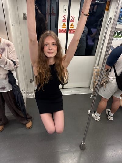 The image shows a young woman with long, wavy blonde hair posing in what appears to be a subway car, possibly in Shanghai's Changning District, China. She is wearing a black dress and holding onto a metal pole for support, with one arm stretched out. Her other arm is also visible, resting near her head. Her expression is a slight smile, and she has a blush on her cheeks, suggesting a lighthearted or playful mood.
In the background, another person's legs are visible, wearing white sneakers and grey shorts, and another person is wearing a light-colored top with a tie-dye pattern and brown pants. There is also a folded black umbrella visible. The setting features the interior of a subway car, with white walls, metal poles, and signage in Chinese characters. One sign clearly reads "爱心专座 COUR", indicating a priority seating area. Another sign, "禁止设备" and "当心夹手", translates to "No equipment" and "Beware of pinched hands," respectively, and "Warning handle pinching," further confirming the subway context. The overall lighting suggests it is daytime, likely inside the subway. The floor of the subway car is a speckled gray.