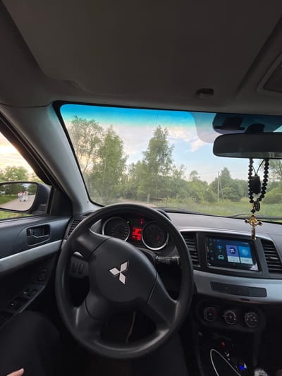 The image is a point-of-view shot from inside a car, looking through the windshield. The steering wheel and dashboard are prominent in the foreground, featuring a Mitsubishi logo on the steering wheel and a digital infotainment screen. Outside the car, a road winds through a tree-lined landscape under a cloudy sky with sunset hues. In the left side mirror, two figures are visible walking away on the road. The car appears to be stationary. The location is identified as Nikolskoye, Russia.
