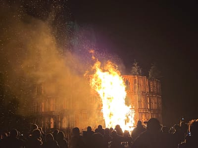 A large bonfire burns intensely, constructed from wooden pallets arranged in stacked tiers. Three small pine trees are visible atop the unburned portion of the structure. A crowd of silhouetted people stands watching the fire, some holding up phones to capture the spectacle. The intense heat creates swirling embers and smoke. The night sky is dark, contrasting with the bright flames.