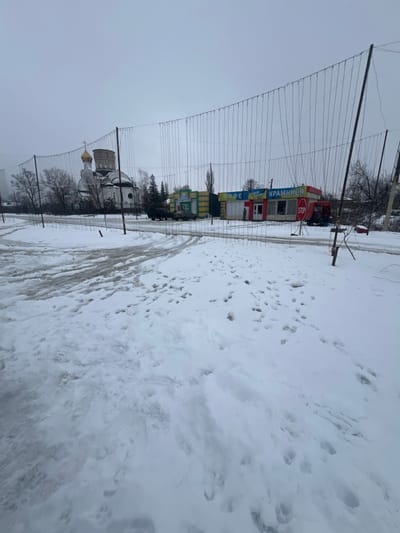 The image depicts a snow-covered street and adjacent area in Staryi Saltiv, Ukraine, under an overcast sky. In the foreground, the ground is covered with white snow, marked by multiple sets of vehicle tracks and numerous footprints. A grid-like structure composed of vertical poles and horizontal wires, from which thinner vertical wires hang, spans across the midground of the scene, partially obscuring the background.
Behind this structure, a dark-colored truck is parked on a snowy area to the left. To the right of the truck, a row of single-story commercial buildings is visible. The leftmost building is yellow and green. Adjacent to it, a red and white building features signs in Cyrillic, reading "КАФЕ" (CAFE) and "КРАМНИЦЯ" (SHOP), with a red hexagonal "STOP" sign positioned near its entrance.
In the background, a white Orthodox church with a prominent golden dome and a cross is visible through and above the wire structure. The church features multi-tiered architectural elements. Bare deciduous trees and some evergreen trees frame the church and commercial buildings. No individuals are visible in the scene.