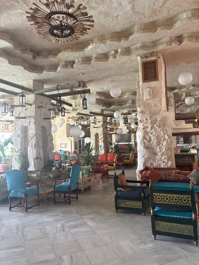 The image shows the interior of a restaurant or cafe with an ornate ceiling and rough-hewn stone pillars. The ceiling features a large, rustic chandelier made of straw-like material and decorative molding that creates a textured, uneven surface. Several white paper lanterns are suspended from the ceiling, along with traditional black lanterns hanging from wooden beams. The pillars are made of textured stone, some with wooden accents.
The seating area includes several distinct furniture sets. In the foreground, two bright teal upholstered chairs with dark wooden frames are positioned around a glass-topped wooden table. Further back, there are low-profile seating areas with ornate green and gold wooden frames, upholstered in various colors including teal and blue, with contrasting throw pillows. To the left, a collection of orange and yellow upholstered chairs and sofas are visible.
Plants are scattered throughout the space, adding a touch of greenery, including potted palms and other foliage. The flooring is a polished white marble with grey veining, laid in large rectangular tiles. The overall ambiance suggests a blend of rustic and exotic decor, possibly inspired by North African or Middle Eastern design. While the image is described as being in Taguermess, Tunisia, no specific identifiable landmarks within that city are visible. The scene is empty of people, with no visible actions or interactions taking place.
