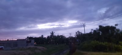 The image shows a landscape in Meunasahteungkudigadong, Indonesia, under a cloudy, twilight sky. In the foreground, there is a field and a small ditch. To the left is a partially constructed building. A line of trees, including palms, stretches across the middle ground, obscuring some structures. Utility poles and wires are visible above the trees. The sky is predominantly filled with gray and purple clouds with a lighter opening near the horizon.