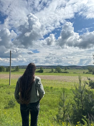 A woman stands in a grassy field on a sunny day. The sky is blue with large, puffy white clouds. Power lines run across the sky, supported by wooden poles. In the background is a green field and a line of trees. The woman has long, brown hair and is wearing a green jacket and patterned leggings. The location is Kremyonki, Russia.