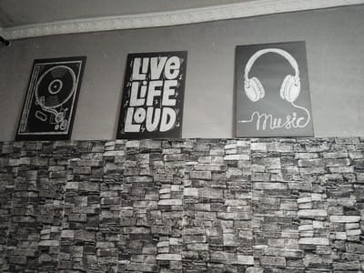 This media file shows three framed posters on a grey wall above a brick-patterned surface. The posters are black with white designs. The left poster features a detailed illustration of a turntable with a vinyl record. The middle poster displays the text "LIVE LIFE LOUD" in large, bold, stylized lettering, with musical notes and lightning bolts as accents. The right poster shows a pair of headphones with a wire connecting to the word "Music" written in a flowing script below. A decorative white molding runs along the top of the wall. The lighting appears to be artificial, and the overall scene suggests an indoor setting, possibly a cafe, lounge, or home entertainment area. The context indicates this location is in Ақадыр, Kazakhstan. There are no people or explicit indications of weather or time of day. The emotion conveyed is one of vibrancy and a love for music.