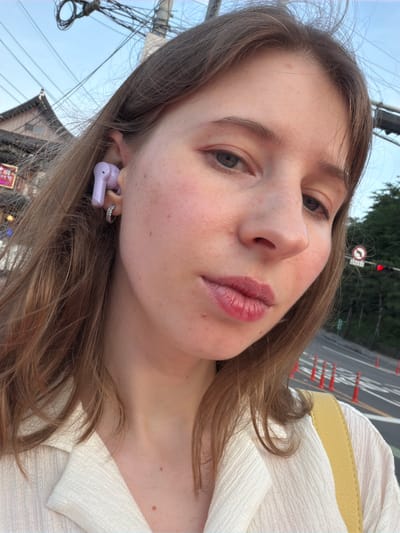 A close-up, slightly angled outdoor selfie features a young woman with shoulder-length brown hair and fair skin. She is wearing a light-colored, textured shirt and a yellow bag strap is visible. She has a small hoop earring in her left ear and a light purple earbud in her left ear. The background shows a street scene with traffic lights, power lines, traditional Japanese-style roofing on buildings, and orange traffic cones. The sky is visible in the upper portion of the image. The location is identified as Pocheon-si, South Korea.