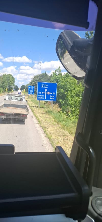 The image shows a road scene in Maslarevo, Bulgaria, on a sunny day. A blue road sign indicates directions to Ruse (3, E83), Svishtov (504), and Resen (504). There is also a smaller blue sign with a white arrow pointing left, indicating directions to Byala (3), and Veliko Tarnovo (2). In the foreground, a white flatbed truck is visible from behind, with the license plate T 7513 BB. Another vehicle is partially visible further down the road. The surrounding environment is characterized by green trees and dry grass. The perspective is from inside a vehicle, likely a truck, with a side mirror and part of the dashboard visible in the frame. The sky is blue with scattered white clouds.