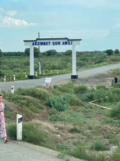 This media file captures a roadside scene in Leninizm, Uzbekistan. The primary subjects are a sign that reads "ARZIMBET QUM AWILI" in large blue lettering, and a smaller sign with the word "MARKET" printed on it, featuring a graphic that resembles an advertisement. The signs are mounted on white posts, forming an archway over a paved road that winds through a landscape of green vegetation and dry, brown earth.
The setting appears to be a rural or semi-rural area. The sky is a pale blue with scattered white clouds, suggesting it's daytime, likely mid-morning or afternoon. The vegetation is varied, with lush green bushes and trees contrasting with patches of dry, sparse growth. The road itself is empty of vehicles, but a person is visible in the background near the road, interacting with some infrastructure. Further down the road, another person is partially visible at the bottom right of the frame, wearing a patterned dress.
Notable details include several smaller white posts along the side of the road, some with black reflective markers. The overall impression is of a functional roadside marker and a small market in a somewhat arid environment. The scene is still, with no discernible activity or strong emotions conveyed. The location cues of "ARZIMBET QUM AWILI" and "MARKET" along with the arid landscape and vegetation are characteristic of the region.