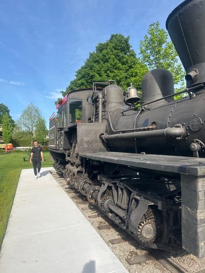 A young man in a black t-shirt and black sweatpants walks on a concrete sidewalk next to a large black steam locomotive. The locomotive is on a train track with gravel and wooden ties. The man is walking away from the camera, towards the front of the train. The locomotive is in the foreground, and the man is in the middle ground. The background shows trees, a blue sky with some clouds, and a building. The image appears to have been taken in Harbor Springs, Michigan, United States of America, at a location that houses historical train equipment. The locomotive has the number "2" painted on its side. A bell is visible on top of the locomotive.