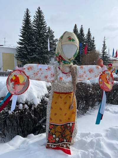 An effigy, styled as a traditional Russian woman, stands upright in a snow-covered public area within the city of Plast, Russia, under an overcast sky. The effigy is dressed in a white headscarf patterned with green leaves and red berries, a white blouse with orange floral patterns, an embroidered vest-like garment, a red necklace, and a yellow apron decorated with red berries and a broad strawberry-patterned border. A braided pigtail hangs from its right side. Its outstretched arms each hold a circular, multi-colored quilted disc, from which red, blue, and white ribbons, emblematic of the Russian flag, extend downwards.
Deep snow covers the ground, obscuring the base of the effigy and nearby low-lying bushes. In the background, numerous tall, snow-dusted evergreen trees rise prominently. To the left, a light-colored building with a snow-covered roof is partially visible. Further back and to the right, several flags, including white, red, and blue, are displayed on poles. Another smaller effigy is partially visible in the distant right background.