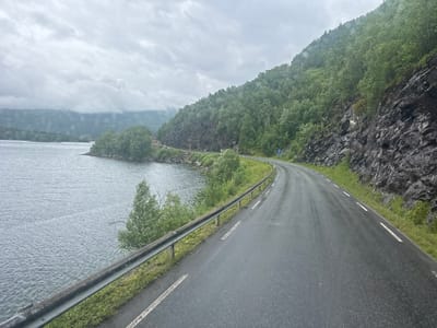 This is a landscape photograph taken on a cloudy day, showing a paved road curving to the left alongside a body of water. The road has white lane markings and a metal guardrail. To the right of the road, a steep, rocky slope covered in lush green trees rises sharply. The water on the left appears calm with small ripples. In the distance, the shoreline continues and is also lined with trees and rocky terrain. The sky is overcast with thick, grey clouds. The image depicts a scenic view of a coastal road, possibly in a mountainous or fjord region of Norway. The location is identified as Bodø, Norway.