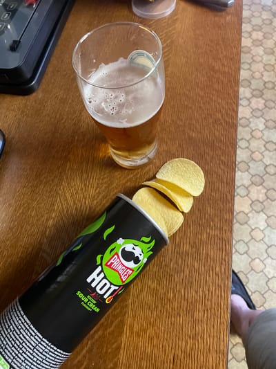 A Pringles can of "Hot Kickin' Sour Cream Flavour" is shown lying on its side on a wooden table in Gerstheim, France. Several potato chips are spilling out of the open top of the can. A glass of beer with a frothy head sits upright on the table near the can. In the background, a black electronic device is partially visible on the left, and a patterned floor is on the right. A person's foot wearing a dark shoe and light-colored pants is visible on the far right of the image.