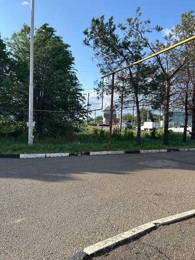 The image shows a section of asphalt road with a white and black curb, bordering a grassy area with trees, likely in a residential or mixed-use area of Zapadny, Russia. A white sign is fixed to a white pole and there is a yellow metallic construction in the frame. Several cars are parked along the road further away. The sky is visible between the trees and is clear with some clouds.