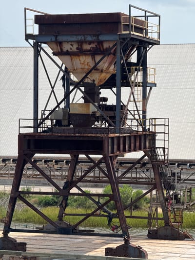 A heavily rusted industrial structure, possibly a conveyor or storage facility, stands on a platform in a body of water. The structure is made of dark metal beams and features a large, trapezoidal hopper at the top, also showing significant rust. There are platforms with yellow railings and stairs attached to the sides of the structure, suggesting it is designed for access and maintenance. The background is dominated by a large, corrugated metal building with a white, reflective surface. Some green vegetation and a chain-link fence are visible behind the structure. The overall scene suggests an industrial area, possibly near a port or manufacturing facility in Brunswick, Georgia, United States of America. No people are visible in the image.