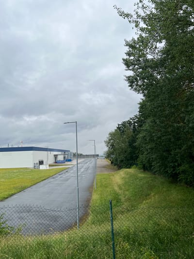 The image depicts an outdoor scene on a cloudy day, likely in the morning or afternoon given the overcast sky. The setting appears to be industrial or commercial, with a large, modern building that has a white facade and dark blue accents. The building has multiple stories and appears to be part of a larger complex, as suggested by the paved area or road leading away from it and the presence of a chain-link fence.
To the left of the building and stretching across the top of the frame is a dense canopy of green trees with lush foliage, suggesting a temperate climate. The ground adjacent to the paved area is covered in vibrant green grass, some of which appears to be somewhat overgrown. The paved area itself looks wet, indicating recent rain or that it is a damp day.
Several tall, slender lampposts are visible along the paved area, spaced at intervals. There are no people visible in the frame. The overall mood of the image is somewhat subdued, likely due to the cloudy weather. The chain-link fence, running along the right side of the image, separates the paved area and grass from the foreground. There is no visible text in the image. The location context provided is Ostrov u Stříbra, Czechia.