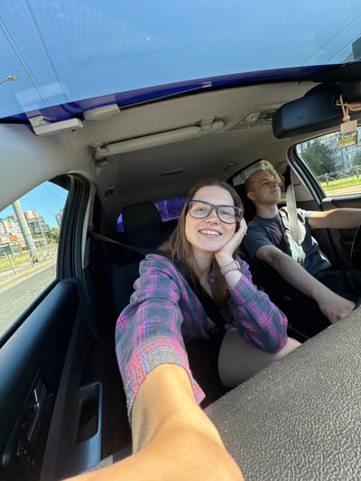 A woman wearing glasses and a pink and blue plaid shirt takes a selfie in the passenger seat of a car, her left hand resting on her cheek. Beside her, a man with a shaved head, wearing a gray t-shirt, drives the car. The car's sunroof is open, revealing a bright blue sky. The vehicle is on a city street in Molodtsovo, Russia. Buildings and trees line the street outside the car's windows, and a fence is visible in the background. The scene appears to be a sunny day, and the overall mood is casual and happy.