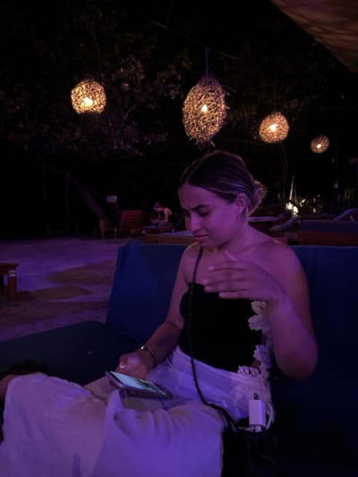 A young woman is sitting on a blue couch, wearing a black strapless top and white pants. She is looking down at a phone in her hand, and her other hand is resting on the couch. There is a white power adapter on the couch next to her. The scene is lit by string lights hanging from the trees in the background. The background is a dark, outdoor setting, with lush green trees. The scene is located in Santa Teresa, Costa Rica, near the beach. There is a small wooden table with a number "38" on it. The coordinates are latitude: 9.632, longitude: -85.158.