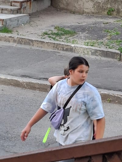 This is a vertical, medium-shot photograph of a young person, likely a girl, standing on a paved outdoor area with grassy patches and concrete steps. The person is wearing a light-colored, tie-dye t-shirt and has dark hair pulled back. A dark strap crosses their chest, supporting a small, purple fanny pack. Their right arm is extended, and they appear to be looking off to the side with a neutral or slightly pensive expression. A bright yellow, tag-like object hangs from the fanny pack. The background shows a rough concrete wall and steps, with some vegetation growing through cracks. The scene appears to be in an urban or suburban setting. The lighting suggests it is daytime, possibly overcast, as there are no harsh shadows. There is no visible text in the image.
