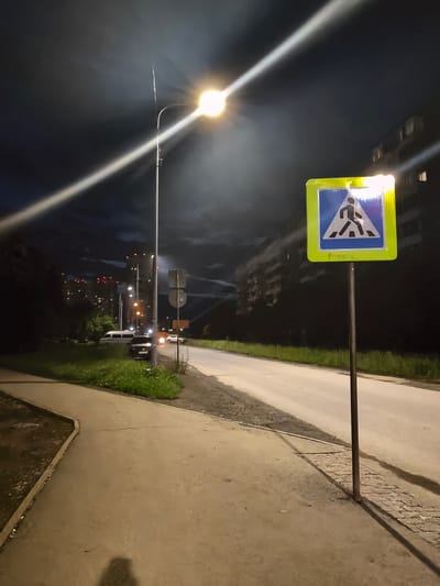 An eye-level, full shot shows a pedestrian crossing sign at night on a sidewalk next to a road. The sign is yellow and blue with a silhouette of a person walking. A street light above casts a strong beam of light across the scene. The sky is dark with clouds. In the background, there are buildings and cars. The sidewalk is concrete, and the road is asphalt. The grass along the side of the road is green. The lighting suggests it is dusk or nighttime, with artificial light sources illuminating the scene.