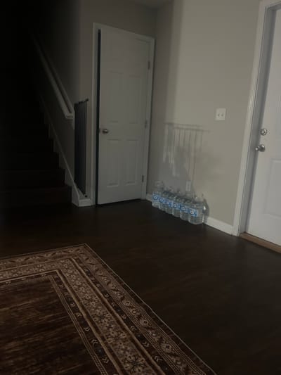 The image depicts an interior scene, likely a hallway or foyer in a residential setting. On the left, a dark staircase ascends into shadow. Adjacent to the stairs, a black metal baby gate is installed. A white door with a silver doorknob is visible in the mid-ground. To the right of this door, a row of plastic water bottles with blue labels are lined up against a light-colored wall. Above the water bottles, a white electrical outlet is mounted on the wall. Further to the right, another white door with two silver doorknobs is partially visible. The floor is covered with dark hardwood and a portion of an oriental-style rug with brown, beige, and blue patterns is in the foreground. The lighting is dim, casting shadows throughout the scene. The location is described as Jacksonville, United States of America.
