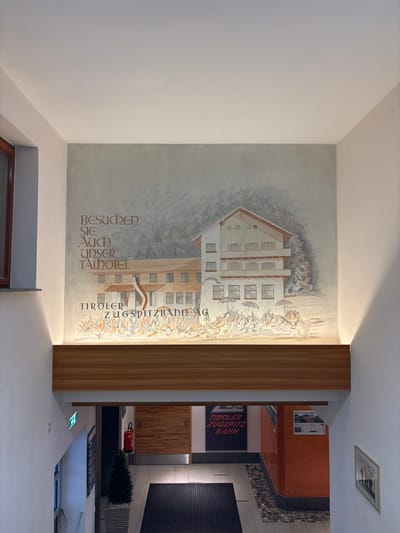 The image shows an interior view of a building, likely a hotel or a public establishment, with a large mural painted on the wall above a wooden beam. The mural depicts a snow-covered alpine scene with a hotel building, trees, and text that reads "Besuchen Sie auch unser Talhotel" and "Tiroler Zugspitzbahn AG". Below the mural, a wooden beam separates the artwork from the lower part of the building, which features a staircase and a hallway with an exit sign, a fire extinguisher, a potted plant, and a framed picture on the wall. The overall impression is one of a welcoming and rustic mountain-themed environment. Based on the text on the mural, this location is likely in Ehrwald, Austria, given its connection to the Tiroler Zugspitzbahn, a cable car railway that goes up the Zugspitze mountain.