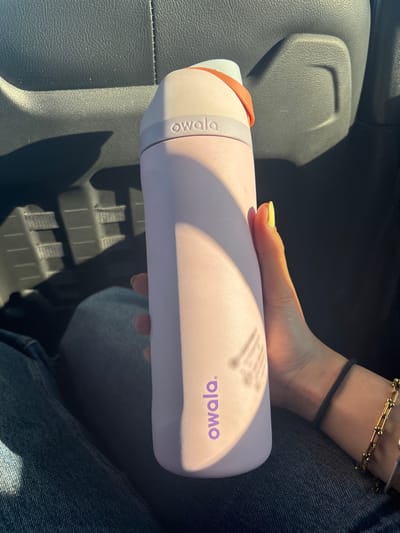 A person is holding a light purple Owala water bottle in their hand. The bottle has a white top and an orange handle. The person is wearing blue jeans and two bracelets on their left wrist, one gold and one black. The scene appears to be inside a vehicle, with a gray leather seat visible in the background. The location is unknown.