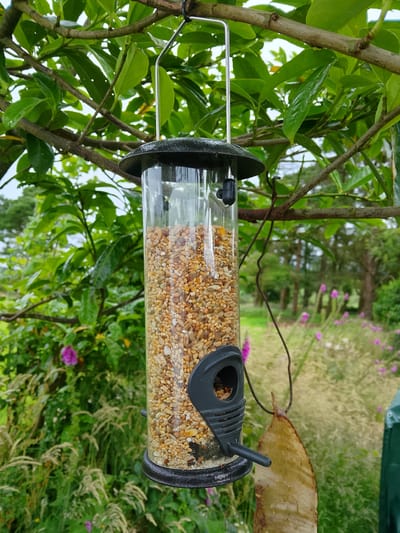 A cylindrical bird feeder, filled with mixed birdseed, hangs from a tree branch in Glounaglough, Ireland. The feeder has a black plastic base and cap, with a black feeding perch extending from the side. A natural, leaf-like object is suspended by a dark string from the bottom of the feeder. The feeder is surrounded by lush green foliage and branches. In the background, a garden scene unfolds with tall grasses, purple flowers, and trees under an overcast sky. The environment appears to be damp, with water droplets visible on the leaves, suggesting recent rainfall.