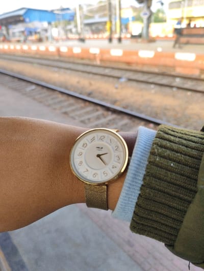 A close-up, first-person view displays a left forearm and wrist, featuring a gold-toned Helix Time analog wristwatch with a white face and a mesh strap. The watch indicates approximately 11:25. The arm, exhibiting a light brown skin tone, is partially covered by a light blue undershirt cuff and a green, ribbed long-sleeved garment. The subject is positioned on a grey concrete surface, likely a platform at a train station in Bardhaman, India. In the blurred background, two sets of railway tracks run diagonally, bordered by an orange-colored wall or barrier. Further in the distance, indistinct blue-roofed station structures, overhead railway lines, and faint human figures are visible on a distant platform, indicating a bustling railway environment.