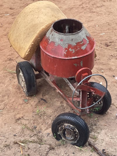 A red, worn concrete mixer rests on sandy ground in Abraka, Nigeria. The mixer exhibits peeling paint and rust. It features a drum, a steering wheel-like mechanism for operation, and three tires. A tan-colored object is positioned near the drum's opening. Small patches of vegetation are scattered near one of the tires.