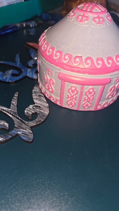 The image shows a close-up of a small, decorative yurt-shaped trinket box with pink and gray detailing, placed on a dark blue surface. To the left of the box, there are two metallic, ornamental shapes with intricate patterns. The background is blurred, showing some other objects, including what appears to be a teal colored object and some blue elements. The overall setting seems to be a tabletop or shelf, possibly in a home or a shop. There is no indication of the city of Leñgir, Kazakhstan in the image itself.