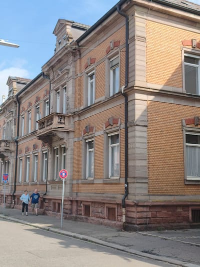 A sunny day in Lörrach, Germany, with a clear blue sky and white clouds. The scene is a street with a sidewalk and an asphalt road. On the sidewalk, a couple is walking towards the camera. The man is wearing a blue t-shirt and shorts, and the woman is in a light blue blouse and dark pants. They are holding hands and appear relaxed.

Behind them, a large, multi-story building with ornate architecture lines the street. The building is constructed with light brown bricks and features sandstone accents around the windows and corners. The windows are tall and have white frames, some with curtains visible. A small balcony with decorative stonework juts out from the second floor. The lower part of the building has a foundation made of rough-hewn reddish-brown stones.

Two street signs are visible on the sidewalk. One is a blue sign with a red border and a red "X" inside a red circle, with the word "TAXIS" below. The other is a circular red and blue sign with arrows indicating no parking or waiting.

The time of day appears to be late morning or early afternoon, given the bright sunlight and the shadows cast on the building and road. The overall impression is a peaceful, everyday scene in a European town.