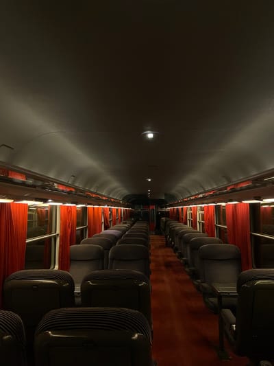 An interior view of a train car, oriented sideways. Rows of seats with striped upholstery are visible on the right. Red curtains hang beside the seats. The left side of the image features a long, dark, curved wall with small lights embedded in it. The perspective emphasizes the length of the train car, stretching into a vanishing point in the distance. The overall tone is subdued and dimly lit.