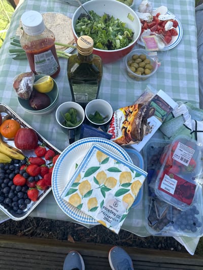 A picnic table in Cloughton, United Kingdom, with a checkered tablecloth. On the table are various food items, including: a bowl of salad, a plate of cherry tomatoes and white cheese, a plate of olives, a plate of fruit, a plate of strawberries, a container of blueberries, a container of grapes, a bottle of ketchup, a bottle of olive oil, and a package of peanut butter puffs. Two bowls with sprigs of mint are on the table. There are also several napkins and a package of crackers on the table. There is a pair of feet in the background.