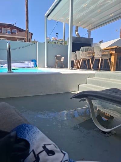 The image depicts an outdoor seating area adjacent to a swimming pool in Nea Iraklitsa, Greece. The scene is bathed in sunlight under a clear blue sky. In the foreground, a portion of a lounge chair with a dark grey cushion is visible, situated on a grey tiled surface partially covered in water. To the left, a metallic railing, possibly belonging to the pool, is partially visible. The swimming pool itself, with its bright blue water, is to the left of the railing. In the middle ground, a light blue wall surrounds the pool area. On top of this wall, a folded white inflatable mattress and a blue pool toy are placed. Further back, a modern outdoor dining area is set up with several light-colored chairs around a wooden table. An awning with integrated lighting extends over this seating area. A barbecue grill is also visible behind the chairs. In the background, a building with brown siding and balconies, along with a wooden utility pole, can be seen. The overall impression is a well-maintained, modern outdoor space designed for relaxation and entertainment.
