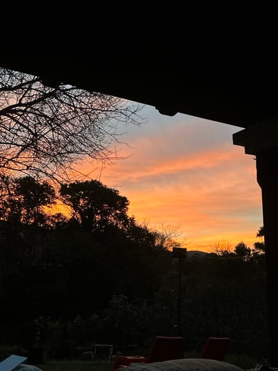 The image captures a sunset viewed from a covered patio in El Rodeo, Argentina. The sky is filled with vibrant orange and pink clouds, indicative of twilight. Silhouetted against the colorful sky are the bare branches of a tree on the left and the dark foliage of trees and bushes forming a dense line across the middle ground. A dark, angular structure, likely part of the patio's roofing or support beams, occupies the top portion of the frame, casting a shadow over the scene. In the foreground, partially visible are two red lounge chairs and the edge of a striped cushion or blanket, suggesting an outdoor seating area. The overall impression is one of a peaceful evening, with the natural beauty of the sunset contrasting with the man-made structures.