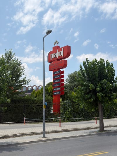 The image shows a tall, red sign with white text that reads "mahal" at the top. Below this, there are smaller sections of text in Turkish, including "Şehrin En Lezzetli", "En Keyifli Ve", and "En Boğaziçi Mekanı". Further down the sign, there are listings for specific items such as "SERPRE KAHVALTI", "FIRINDAN PIZZALAR", "LEZZETLI YEMEKLER", "GÜVÖN ALANI", "DÖNEY & NARGILE", and "MAÇ KEYFI". The sign is mounted on a pole and is located outdoors. To the left of the sign is a streetlight. In the background, there are trees, bushes, and some arched structures visible, suggesting a commercial area or restaurant. The ground in front of the sign is paved, and there are some orange and white safety cones and tape. The sky is blue with some white clouds. The scene appears to be in Denizli, Turkey, as indicated by the Turkish text on the sign.
