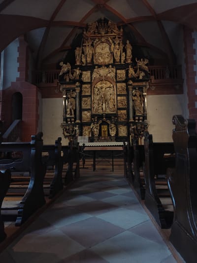 This image depicts the interior of a historic church, located in Aschaffenburg, Germany. The view is from the rear of the nave, looking forward along a central aisle towards the main altar and altarpiece.
In the foreground, dark, ornately carved wooden pews line both sides of the aisle. The floor is paved with a checkerboard pattern of light grey and darker reddish-brown square tiles. No individuals are present in the pews or aisle.
The midground is dominated by a large, multi-tiered altarpiece, constructed from dark wood or stone, positioned centrally. The altarpiece is intricately carved, featuring numerous sculpted figures and relief panels. A prominent crucifixion scene is visible in the central upper section. Below this, numerous figures of saints and angels, along with narrative scenes, are arranged in various niches and panels. A small, golden tabernacle-like structure is visible at the very bottom center of the altarpiece, resting on an altar table covered by a white, lace-edged cloth.
The background reveals the church's architectural elements. The walls are predominantly light-colored, with structural elements composed of dark red sandstone or brick, forming robust pillars and arches on the left and right. On the far left, a high, dark arched recess or doorway is visible within the red stone wall, accompanied by a short flight of stairs. Above, the ceiling is vaulted, characterized by dark wooden ribs supporting lighter, likely plastered, infill panels. Wooden balconies with railings are visible high above the nave on both the left and right sides. The scene is devoid of people, activity, or interactions. The lighting is dim and atmospheric, casting shadows that emphasize the architectural and sculptural details.