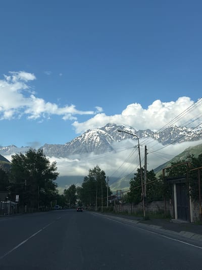The image depicts a scenic view of a road alongside a majestic, snow-capped mountain range. The sky is a vibrant blue, dotted with fluffy white clouds that partially obscure the peaks. Power lines crisscross the frame, indicating the presence of human infrastructure near the natural landscape. On the right side of the road, trees with lush green foliage line the way, and some buildings are visible, including a structure with a metal gate. A few cars are driving on the road, suggesting it is a route for transportation. The perspective suggests the photo was taken from a moving vehicle. The bright sky and the clear visibility of the mountains suggest it is daytime, likely late morning or afternoon, and the weather appears to be fair. The overall impression is one of natural beauty enhanced by the accessibility of a road. A sign with "HOTEL" is visible on the left side of the road.