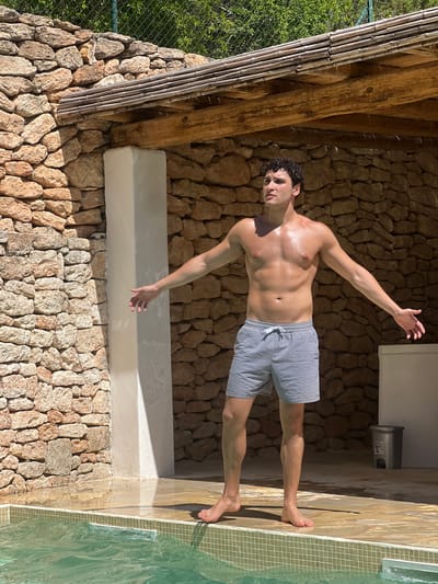 A young man with curly dark hair is posing on the edge of a swimming pool. He is leaning back, with his left arm extended upwards and his right arm bent at the elbow, hand resting on his hip. He is wearing blue and white striped swim trunks. The wall behind him is constructed of natural stone. To his left, there is a wooden beam supported by bamboo poles, suggesting an outdoor shelter or pergola. Sunlight casts shadows on the stone wall, indicating it is daytime. The water in the pool is clear and green, with small square tiles lining the edge. The overall scene suggests a relaxed, vacation-like atmosphere. There are no visible emotions expressed, beyond a casual pose. No text is visible in the image.