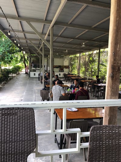 This image depicts an outdoor dining area with a corrugated metal roof supported by metal beams and a large wooden pillar on the right. String lights are visible along the edge of the roof. Several tables and chairs are arranged under the covered area. In the foreground, a brown wicker chair is partially visible. In the midground, a group of men are seated at tables, some appearing to be eating or conversing. One man in a black and white patterned shirt is facing away from the camera. To the left, a concrete walkway extends into the background, lined with green bushes and trees. A woman in a pink hijab and patterned clothing is walking away from the camera down this walkway. The background shows dense foliage and trees, suggesting a natural or park-like setting. The scene appears to be a casual dining establishment in Keudealue Ie Puteh, Indonesia.