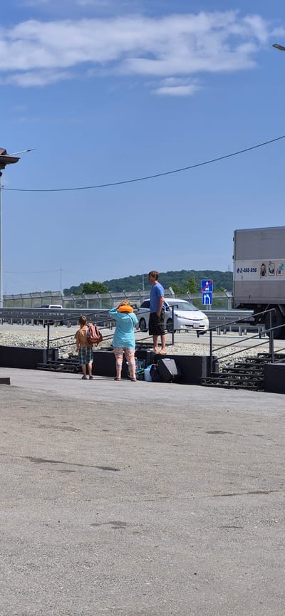 In Taezhnyi, Russia, under a clear blue sky with scattered clouds, three individuals stand near a road. A young boy, wearing a tan shirt and plaid shorts, is seen from behind, carrying a brown backpack. Beside him, a person wearing a turquoise shirt and patterned leggings is adjusting an orange neck pillow. To their right, a man in a blue t-shirt and dark shorts stands with his hands in his pockets, facing away from the camera. Luggage, including two suitcases and a duffel bag, is placed on the ground near the individuals. In the background, a white car and a truck are visible on the road. A guardrail separates the road from a paved area where the people are standing. Lush green trees and hills form the distant landscape.
