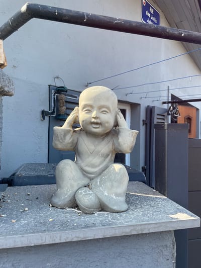 A close-up, rotated view shows a light gray stone statue of a baby in a relaxed pose. The statue is positioned on a textured gray stone surface, with a dark gray structure behind it. The baby statue has its legs bent and its arms raised, with hands near its head. The baby's face is slightly turned, with its mouth open as if speaking or laughing. The statue has a weathered appearance with visible cracks and debris on its surface.
In the background, a light blue sign with white text is visible on a dark pole. The text on the sign reads "RUE des Juifs". Above this, a building with a dark awning and windows can be seen. A black pipe runs diagonally across the left side of the image. The scene appears to be outdoors, possibly on a balcony or ledge, with the sky suggested by the lighting. The time of day is not explicitly clear, but the lighting suggests it might be daytime. The weather appears to be clear, with no visible rain or clouds. The location context is Gerstheim, France, with the street name providing a specific clue to the location.