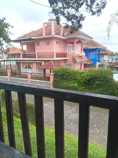 The scene depicts a residential area in Pô Kô, Vietnam, viewed from an elevated perspective on a cloudy day. In the immediate foreground, a dark-colored wooden or metal railing, likely part of a balcony, runs horizontally, partially obscuring the lower section of the view. Below the railing, a green grassy slope leads down to a light-colored, possibly unpaved, street or path.

Beyond the path, a low fence with a light pink stucco base and black railings encloses properties. Behind this fence, a dense green hedge runs in front of a prominent, three-story residential building. This main building is painted a light salmon pink and features a reddish-brown tiled roof. A blue and white water tank is visibly mounted on a raised support structure on the roof. The house has multiple windows, some with white frames and others utilizing glass blocks, along with balconies featuring dark railings and white architectural accents. To the left of the pink house's ground level, a small, octagonal gazebo with a tiled roof is visible.

To the right of the pink residence, a two-story house painted a vibrant blue with a similar tiled roof is partially visible. Further to the right, another house appears green. The background includes scattered trees and dense green foliage, with an overcast, light gray sky overhead. No individuals or specific actions are discernible in the image.
